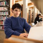 young-man-working-on-laptop-sitting-at-desk-in-busy-public-library