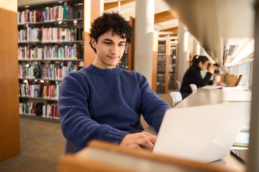 young-man-working-on-laptop-sitting-at-desk-in-busy-public-library